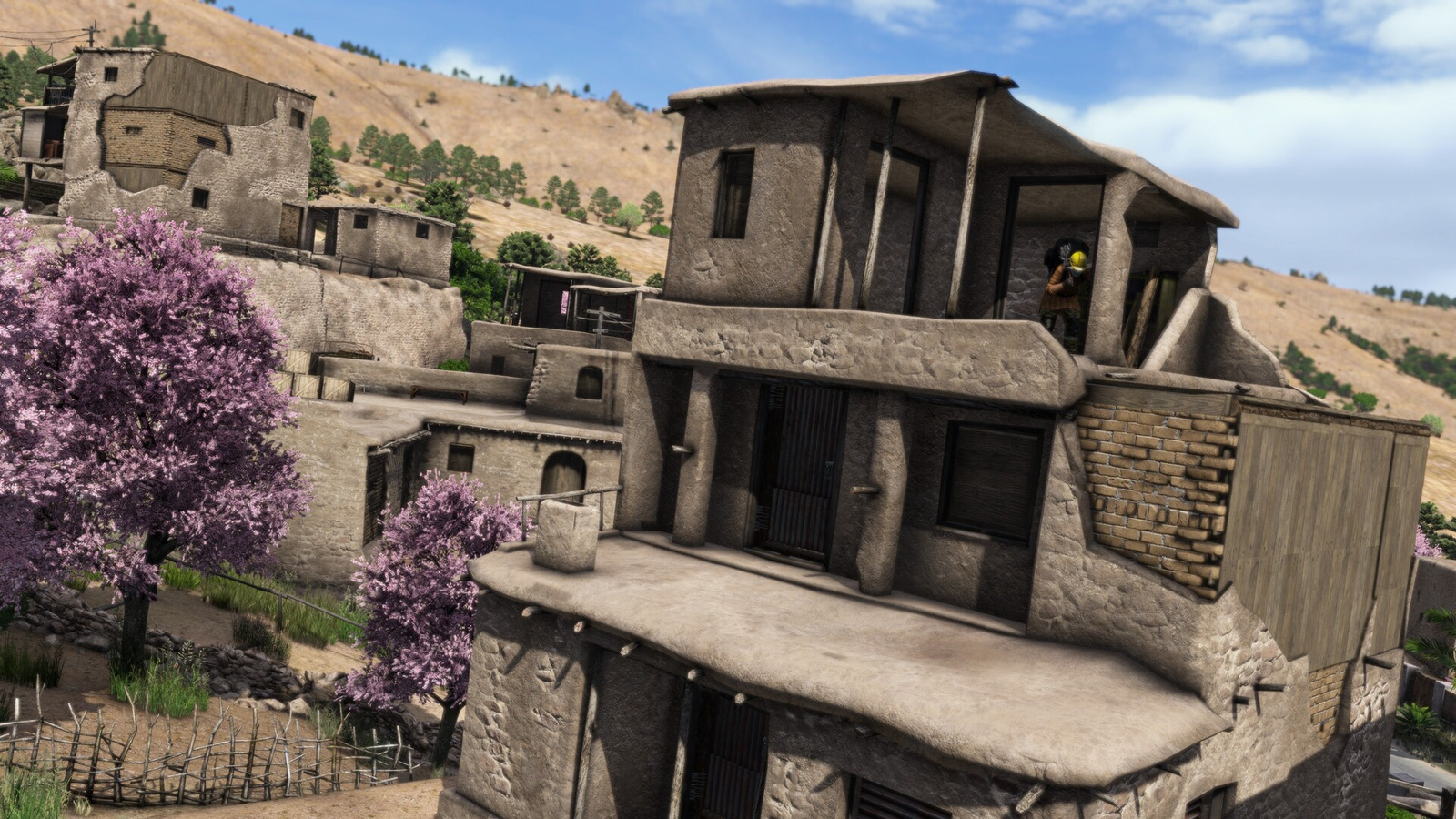 DayZ Badlandsの住宅外観。石や土の質感を持つ多層の建物と、周囲に咲く木々。